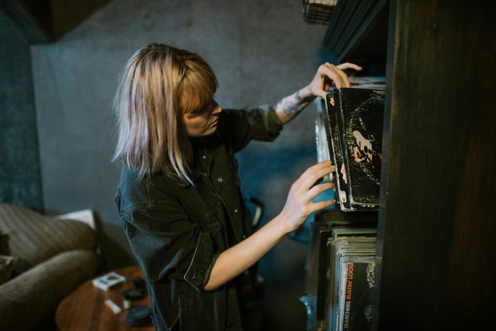 Une personne aux cheveux mi-longs et tatoués, vêtue dune chemise sombre, est immergée dans la musique de rituel tout en regardant une étagère de disques vinyles dans une pièce confortable avec un canapé et une table en bois.