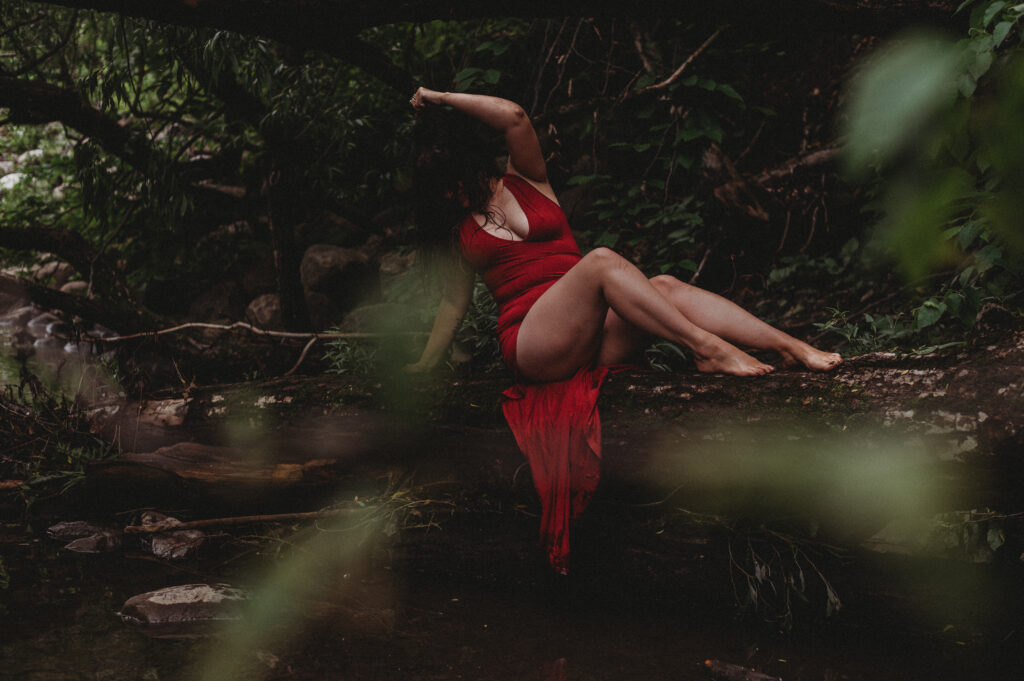 Une personne vêtue dune robe rouge fluide est assise sur une bûche entourée dune végétation luxuriante et dun petit ruisseau. Elle est pieds nus, un bras levé et les jambes croisées, évoquant une scène enchanteresse à la fois sereine et magique, comme laura dune sorcière sexuelle secrète cachée dans létreinte de la nature.